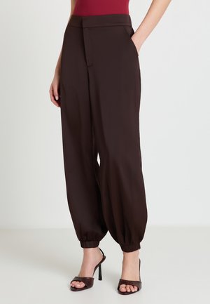 Donna che indossa pantaloni a vita alta marrone scuro a gamba affusolata con polsini elastici e sandali con tacco alto e punta aperta, in piedi con le mani nelle tasche.