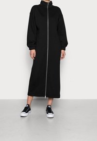 Sort, lynlås sweatshirtkjole med høj krave og puffede ærmer, som har en lang silhuet og elastiske manchetter. Båret med sorte sneakers.