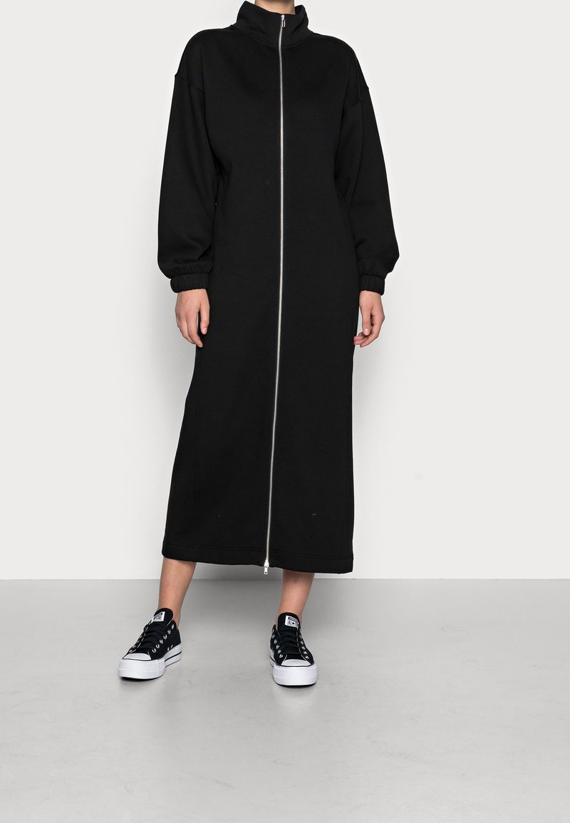 Sort, lynlås sweatshirtkjole med høj krave og puffede ærmer, som har en lang silhuet og elastiske manchetter. Båret med sorte sneakers.