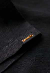 Zwarte stof met een gestructureerde afwerking, voorzien van een opvallend oranje "SUPERDRY" label dat in de rand is genaaid. Eenvoudig, strak ontwerp.