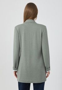 Blazer gris sur mesure avec une coupe légèrement surdimensionnée, présentant un col cranté et des détails de manches retroussées, fabriqué dans un tissu au toucher lisse.