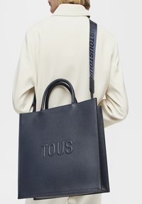 Granatowa skórzana torba tote z wytłoczonym logo "TOUS", podwójnymi uchwytami i regulowanym paskiem na ramię z nazwą marki, wyróżniająca się gładką teksturą.