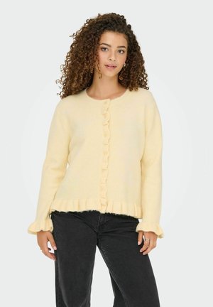 Gilet en maille jaune avec un devant froncé, un col rond et des poignets froncés, associé à un jean noir. Texture douce et fermeture à boutons.