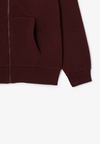 Manche et poche kangourou d'une veste zippée bordeaux avec poignets et bas côtelés sur fond blanc.