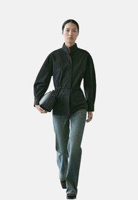 Schwarzes langärmliges Hemd mit hohem Kragen und Knöpfen vorne, kombiniert mit hell gewaschenen, ausgestellten Jeans und einer kleinen schwarzen Tasche. Glatte Textur.