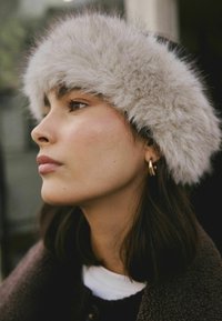 Faux fur pandebånd i lys grå med en fluffy tekstur. Model iført en brun jakke og guldørering, vendt mod venstre.