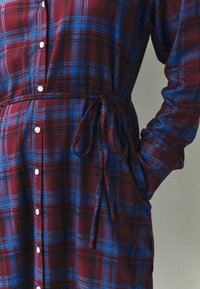Kjole med tern i burgunder og blå, med bånd i taljen, knapper foran og lange ærmer. Fremstillet af blødt stof med et ternet mønster.