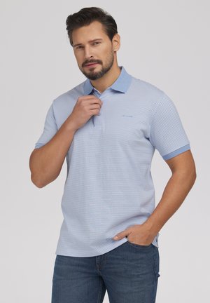 Mężczyzna z brodą w jasnoniebieskiej paskowanej koszulce polo i dżinsach, poprawiający kołnierzyk jedną ręką, a drugą opierający się w kieszeni.