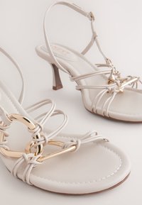 Sandales à talons hauts beiges avec lanières, ornées de ferrures dorées brillantes, présentant des lanières entrelacées et une semelle lisse et rembourrée.