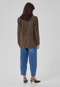 Blazer din suede maro, cu o croială ajustată, ventrală posterioară și rever în formă de ciucure, purtat cu blugi largi de culoare albastră deschisă și tocuri negre cu slingback.