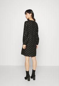 Vestido preto com bolinhas prateadas, mangas longas, decote em V e cintura cingida. Usado com botas pretas de cano curto, exibindo um caimento descontraído.