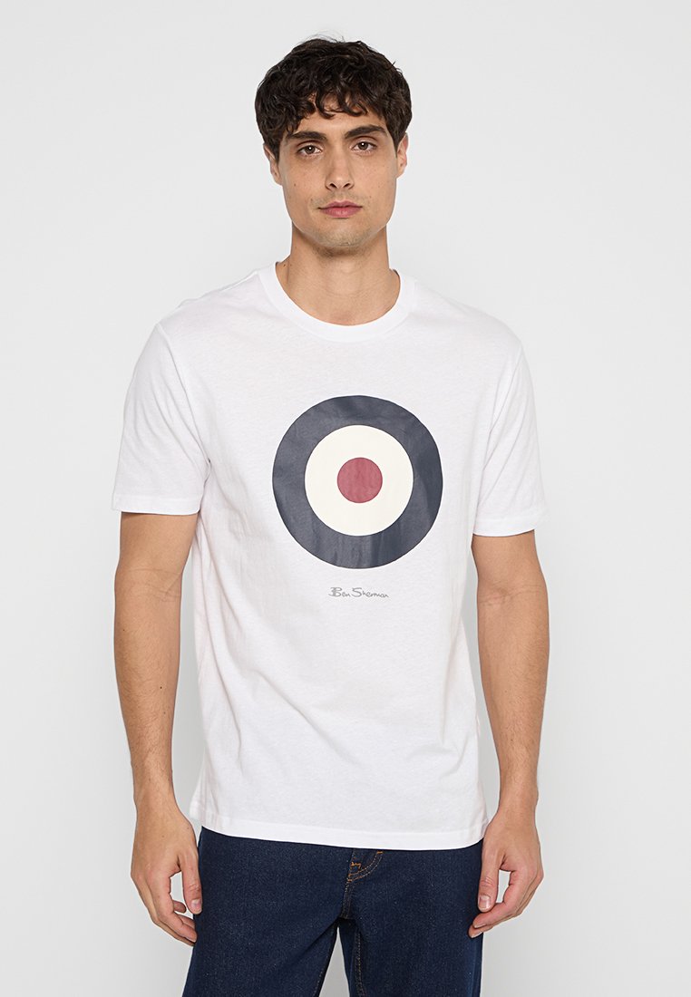 Man in een wit T-shirt met een marineblauw, wit en rood doelontwerp en donkerblauwe jeans, staand tegen een effen achtergrond.