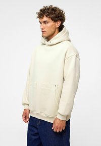 Beige hoodie met een voorkoord, trekkoordcapuchon en een ontspannen pasvorm. Zacht materiaal en zichtbare stiksel details op de naden. Gecombineerd met donkere spijkerbroeken.