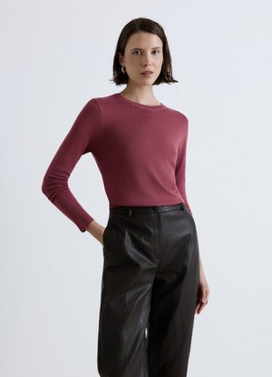 Donna con capelli corti e scuri che indossa una maglia a maniche lunghe color malva e pantaloni in pelle neri a vita alta su uno sfondo chiaro e uniforme.