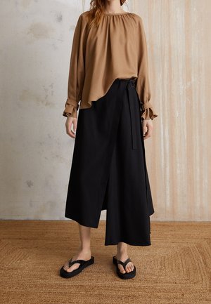 Kvinde iført en løs brun bluse med lange ærmer, sort omslagsnederdel i midi-længde og sorte klip-klap-sandaler, stående på en tekstureret måtte.