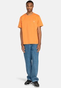 T-shirt en coton orange avec un col rond et des manches courtes, associé à un jean en denim bleu à coupe décontractée et des baskets noires.