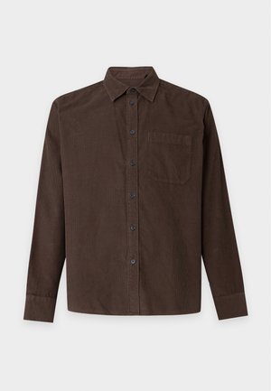 Homme portant une chemise marron foncé à boutons sur un t-shirt blanc, pantalon marron foncé et baskets blanches, debout avec une main dans la poche.