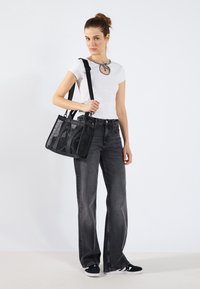 Bolsa tote preta transparente com alças duplas, design estruturado e costuras distintas. O modelo usa uma blusa branca ajustada e jeans cinzentos de perna larga.