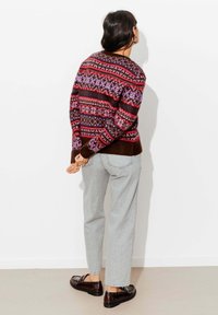 Pull en tricot multicolore avec des motifs géométriques en rouge, rose et violet. Associé à un pantalon gris clair et des chaussures marron.