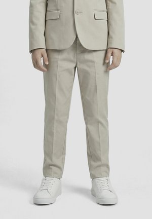 Hombre vestido con pantalones de traje beige a medida y blazer a juego, con zapatillas deportivas blancas con cordones, de pie contra un fondo liso.