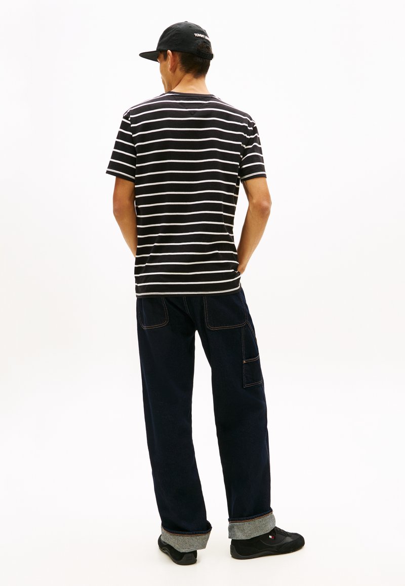 Homme portant un t-shirt à rayures noires et blanches, un pantalon cargo sombre aux revers remontés, des baskets noires et une casquette de baseball noire, vu de dos.