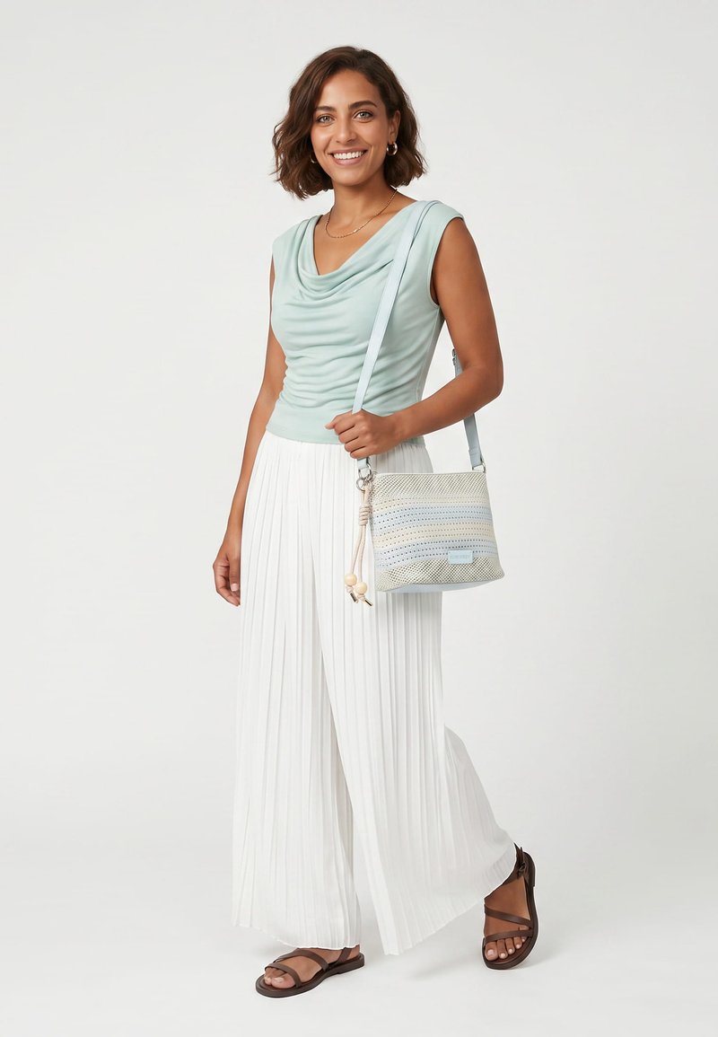 Mujer sonriente que lleva una camiseta sin mangas de color azul claro, pantalones blancos plisados de pierna ancha, sandalias marrones y lleva un bolso beige cruzado con rayas azules.