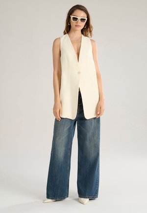 Cream sleeveless vest with a single button, paired with light blue wide-leg denim jeans and pointed white heels. Modern sunglasses complete the outfit.