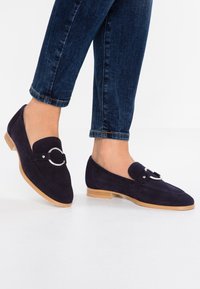 Persona che indossa mocassini in camoscio blu scuro con dettagli in anello argentato, abbinati a jeans blu scuro arrotolati su un pavimento bianco.