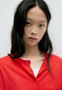 Jeune femme aux longs cheveux noirs et à l'expression neutre, portant un cardigan rouge par-dessus un haut blanc sur un fond uni.