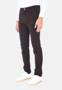 Jeans slim fit marroni scuri con cuciture visibili, tasche frontali e chiusura a bottone singolo, abbinati a sneaker bianche basse.