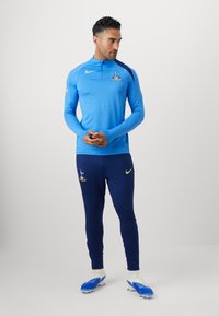 Blå langærmet atletisk trøje med høj krave og logo, parret med marineblå bukser med logo og gul detalje. Hvide og blå fodboldstøvler.