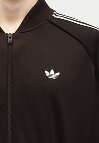 Κοντινό πλάνο ατόμου που φοράει μαύρο μπουφάν με φερμουάρ, με λευκές ρίγες στους ώμους και ένα μικρό λευκό λογότυπο Adidas στο στήθος.