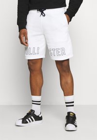 Vita svettshorts med "HOLLISTER" tryckt i fet svart text. Justerbar marinblå dragsko och sidofickor. Bärs med svarta Adidas-sneakers.
