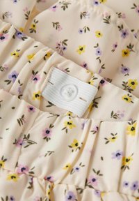 Tissu à motif floral avec de petites fleurs roses, jaunes et violettes sur un fond beige clair ; comprend un bandeau élastique blanc et une fermeture à bouton.
