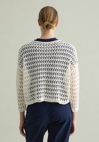 Maglione bianco all'uncinetto con un design morbido, caratterizzato da un colletto blu scuro e maniche lunghe. La texture è a maglia aperta con un motivo geometrico.