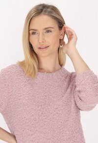 Heller rosa strukturierter Pullover mit rundem Ausschnitt und lockeren Ärmeln. Das Model trägt einen goldenen Ohrstecker mit einem durchsichtigen Akzent.