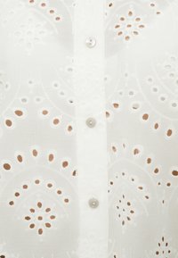 Blouse blanche brodée présentant des découpes complexes, des motifs floraux subtils et des boutons ronds le long de la patte de boutonnage avant.