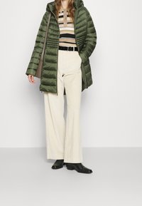 Kvinde iført olivengrøn, kviltet frakke, stribet beige sweater, cremede bredbundede bukser, sorte støvler og bærende på en taupe skuldertaske.