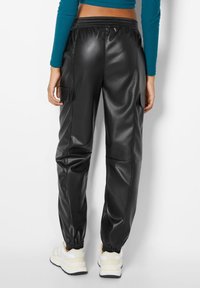 Černé joggers z umělé kůže s elastickým pasem, bočními kapsami na cargo a elastickými manžetami. Model má na sobě bílé a béžové tenisky.