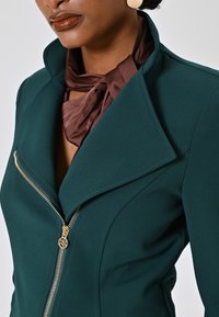 Donkergroene op maat gemaakte blazer met een scherpe kraag en gouden ritsdetails, gecombineerd met een bruine satijnen sjaal om de hals.