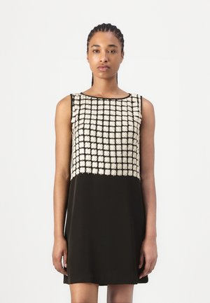 Mujer con el pelo trenzado que lleva un vestido negro sin mangas con un patrón de rejilla floral blanco en la parte superior, de pie frente a un fondo liso.
