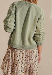 Frau in einem hellgrünen, grob gestrickten Pullover mit Puffärmeln und einem beige-blumigen Rock, von hinten gezeigt.