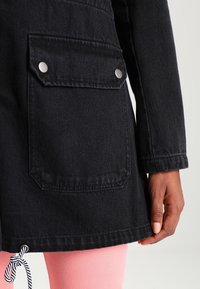 Mustat farkkutakki, jossa on etupuolella läppätasku, hopeiset napit ja säätönyöri helmassa. Sujuva pinta, rento muotoilu.