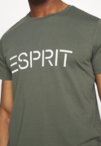 Olivgrünes Baumwoll-T-Shirt mit kurzen Ärmeln, mit einem auffälligen weißen Druck, der "ESPRIT" auf der Brust zeigt. Glatte Textur.