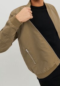Tan bomber jacket with a smooth texture, ribbed cuffs, and a silver zipper. Features a zippered pocket on the side. Worn over a black shirt.