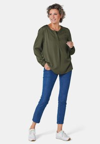 Olivgrünes Langarmbluse mit gespaltenem Ausschnitt, kombiniert mit blauen Skinny Jeans und weißen Sneakers, an einem Modell, das steht und lächelt.