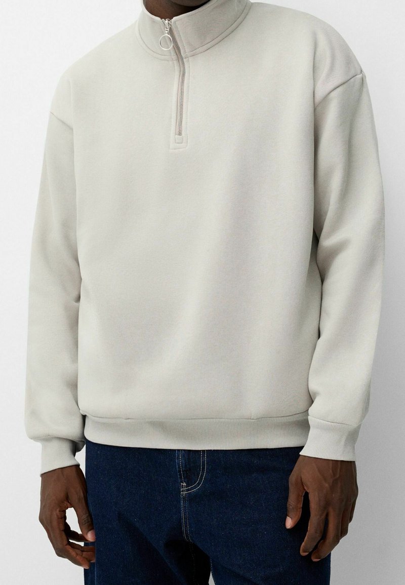 Personne portant un pull en polaire beige clair avec une courte fermeture éclair et un anneau au niveau du col, associé à un jean bleu foncé.