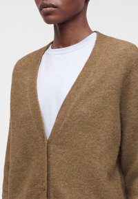 Personne portant un cardigan doux brun pelucheux sur un t-shirt blanc à col rond, montré du cou jusqu'au milieu du torse sur un fond uni.