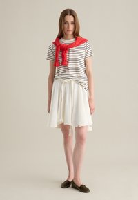 Camiseta de manga corta a rayas, suéter de punto rojo atado al cuello, falda blanca con cintura trenzada y borlas, zapatos verdes tipo slip-on, fondo neutro.