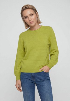 Frau mit kurzen blonden Haaren, die einen limettengrünen, gerippten Pullover und blaue Jeans trägt, steht mit einer Hand in der Tasche vor einem grauen Hintergrund.